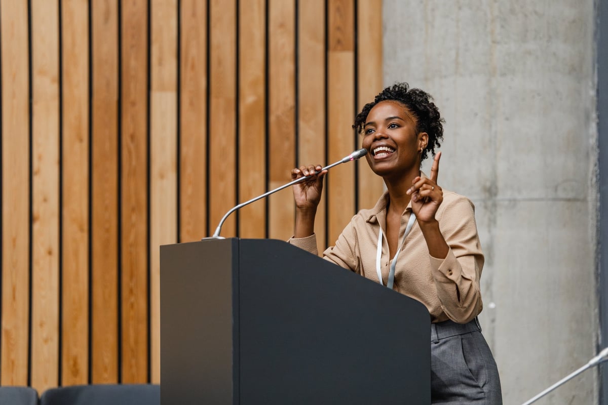 Young professional woman delivering a confident, engaging presentation at a conference podium, smiling and gesturing while speaking into a microphone.