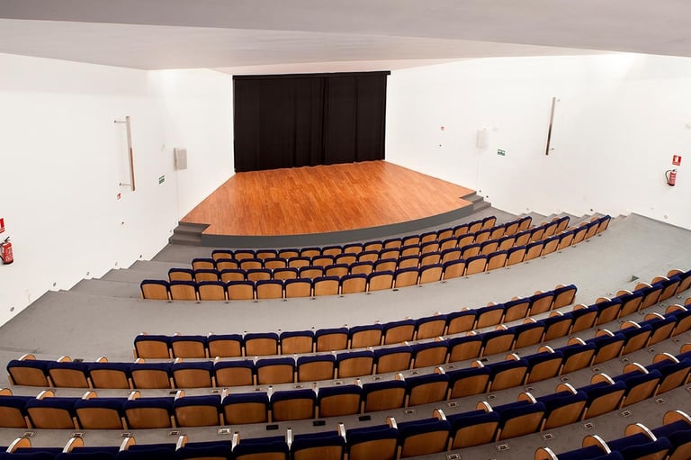 Theater auditorium with rows of orange and black seats facing a wooden stage with dark curtains